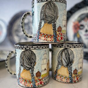 May include: Stacked ceramic mugs with a hand-painted design. Each mug features a woman with braided hair, facing away, with a church and floral details. The mugs have a black and white patterned rim and handle.