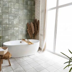 May include: A white oval freestanding bathtub with a wooden tray across the center, positioned against a wall of square sage green tiles. A wooden stool with a woven basket and white towels sits nearby, with dried foliage in the corner.