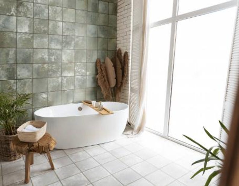 May include: A white oval freestanding bathtub with a wooden tray across the center, positioned against a wall of square sage green tiles. A wooden stool with a woven basket and white towels sits nearby, with dried foliage in the corner.