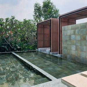 May include: An outdoor water feature with a shallow pool lined with square green tiles. Two wooden pergola structures with vertical slats stand behind the pool, framing a central wall covered in matching square green stone tiles.