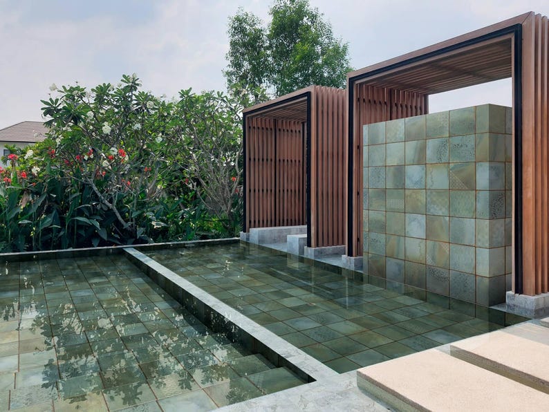 May include: An outdoor water feature with a shallow pool lined with square green tiles. Two wooden pergola structures with vertical slats stand behind the pool, framing a central wall covered in matching square green stone tiles.
