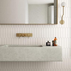 May include: A modern bathroom with a light gray concrete sink and a gold faucet. The walls are covered in white, elongated tile. A mirror and a gold sconce are also visible.