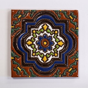 May include: Square ceramic tile with a detailed, colorful design. The tile features a central white floral pattern with a blue center, surrounded by a blue border and a brown outline. The corners have green and yellow leaf-like accents, set against a reddish-brown background.