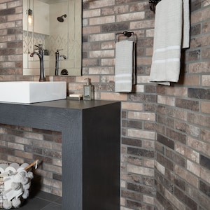 May include: A bathroom vanity with a white sink and a black countertop. The vanity is in front of a brick wall with a mirror and towel racks. There are towels hanging on the racks.