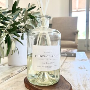 May include: A clear glass bottle diffuser with a wooden base. The bottle is filled with a light yellow liquid and has a label that reads "SCENT NO. 03 POLIGNANO A MARE CLARY SAGE & SEA SALT Casa Mare Collection LUXURY AROMATHERAPY REED DIFFUSER 700ml / 24.0 fl. oz."