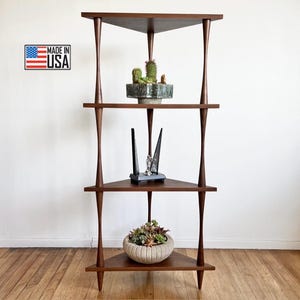 May include: A three-tiered corner shelf unit made of dark wood with a triangular shape. The shelf has three shelves and six legs. The shelves are empty except for a small potted plant on the top shelf, a decorative object on the middle shelf, and a larger potted plant on the bottom shelf. The shelf is made in the USA.