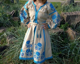 May include: A beige knee-length dress featuring vibrant blue floral embroidery on the sleeves and skirt. The garment includes a V-neckline with blue tassel ties, long puffed sleeves, a matching waist tie belt, and a blue pom-pom trim along the hem.