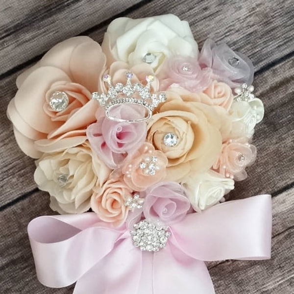 Pode incluir: Um buquê de flores rosa e pêssego com uma coroa de strass e uma fita de cetim rosa. O buquê é perfeito para um casamento, baile de formatura ou uma ocasião especial.