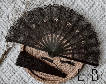 May include: Black lace hand fan with a floral design, open and closed. The open fan displays intricate details, while the closed fan is compact. The fans are on a woven surface. The image also includes the text "Lassos Boutique".