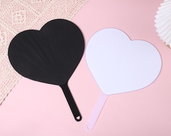 May include: Two heart-shaped hand fans with flat surfaces and long handles. One fan is solid black with a black handle, and the other fan is solid white with a light pink handle. Both fans feature a small circular hole at the end of the handle.