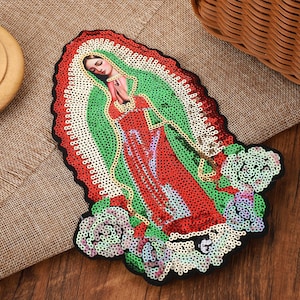 May include: A decorative patch featuring a sequined depiction of the Virgin of Guadalupe. The design includes a figure in a red dress and green cloak, surrounded by a white sequined aura and light-colored floral accents at the base.
