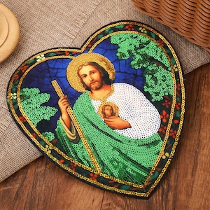 May include: A heart-shaped patch featuring a sequined depiction of a figure with a halo, wearing a white and green robe. The design includes a blue background with green foliage, a gold border, and a small circular emblem on the chest area.