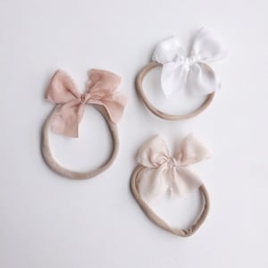 May include: Three nude-coloured elastic headbands with fabric bows in different shades of pink and white. The bows are made of a soft, textured fabric.