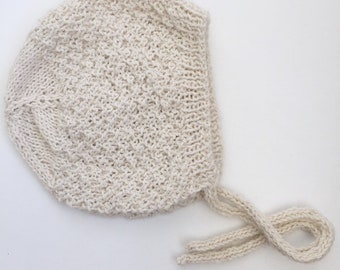 May include: A cream-colored knitted baby bonnet with a long tie. The bonnet is made of a soft, textured yarn.