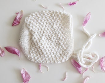 May include: A white knitted baby bonnet with a textured pattern and ties. The bonnet is surrounded by pink flower petals.