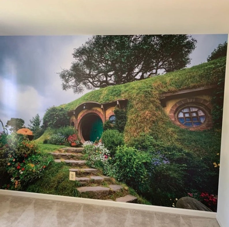 Pode incluir: Uma porta verde com um arco redondo leva a uma casa hobbit com um telhado gramado. A casa &eacute; cercada por vegeta&ccedil;&atilde;o exuberante e flores.