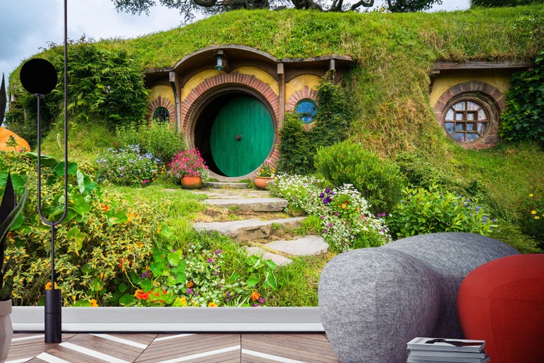 Pode incluir: Uma porta verde com uma moldura redonda &eacute; a entrada para uma toca de hobbit, uma pequena casa redonda constru&iacute;da em uma colina gramada. A casa tem uma fachada de tijolo e uma pequena janela. A cena &eacute; cercada por vegeta&ccedil;&atilde;o exuberante e flores.