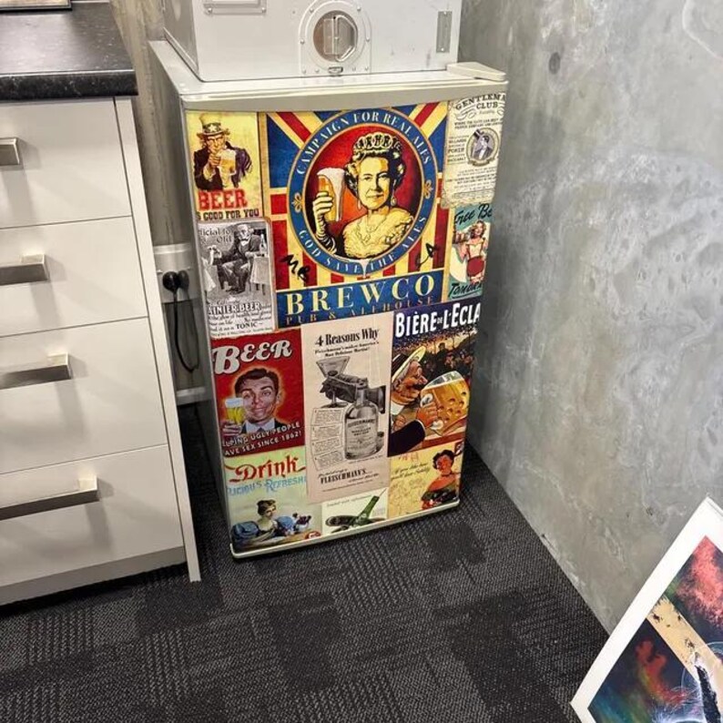 Puede incluir: Un refrigerador compacto cubierto con un collage de anuncios de estilo vintage con tem&aacute;tica de cerveza y pubs. La parte frontal presenta varias ilustraciones, incluido un retrato de una reina y textos que dicen Campaign for Real Ales, Brewco y Beer.