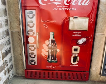 May include: A red Coca-Cola vending machine featuring the text Drink Coca-Cola In Bottles, Serve Yourself, Delicious and Refreshing, Coin Return, Bottle Opener, and Ice Cold. The machine displays a graphic of a hand holding a glass bottle.