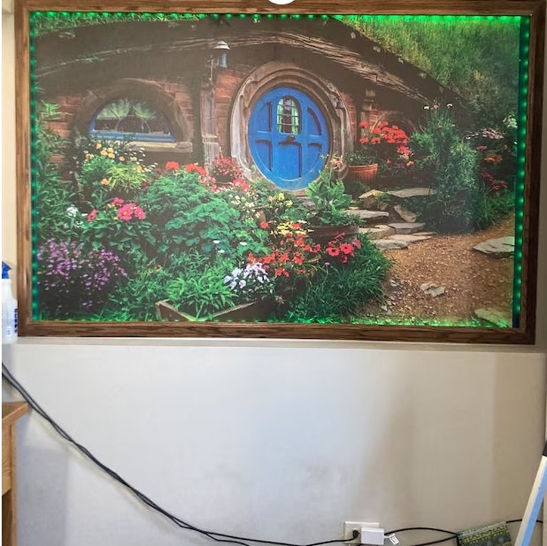 Pode incluir: Uma impress&atilde;o emoldurada de uma casa hobbit com uma porta azul e um jardim com flores e vegeta&ccedil;&atilde;o. A casa &eacute; cercada por uma moldura de madeira com uma borda verde.