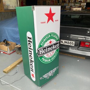 May include: A green and white Heineken refrigerator with a red star and the words "TRADE MARK" on the top. The refrigerator has a Heineken logo with the words "Heineken Pilsener" and "Premium Quality".