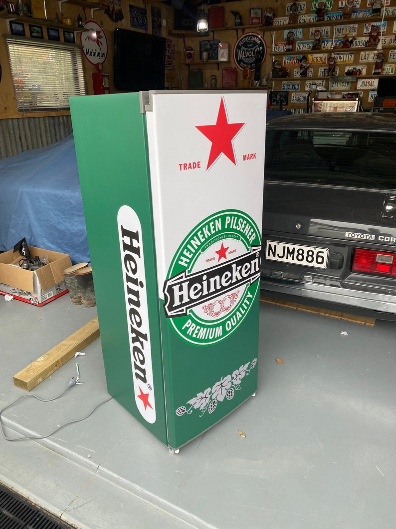 May include: A green and white Heineken refrigerator with a red star and the words "TRADE MARK" on the top. The refrigerator has a Heineken logo with the words "Heineken Pilsener" and "Premium Quality".