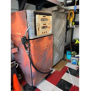 Può includere: Una pompa di benzina in stile vintage con una finitura arancione e rugginosa. L'unità presenta un pannello centrale con contatori numerici meccanici etichettati Total Sale e Gallons, e un tubo flessibile nero con una pistola metallica.