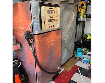 Puede incluir: Una bomba de combustible de estilo vintage con un acabado naranja y oxidado. La unidad cuenta con un panel de visualización central con contadores numéricos mecánicos etiquetados como Total Sale y Gallons, y una manguera negra con boquilla.