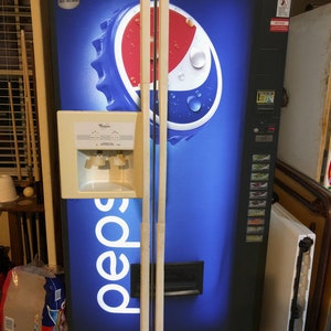 Può includere: Un alto distributore automatico nero a marchio Pepsi con una grande grafica di un tappo di bottiglia blu, rosso e bianco. L'apparecchio include un distributore d'acqua beige sulla porta sinistra, un pannello di selezione verticale e il testo CALORIES COUNT.