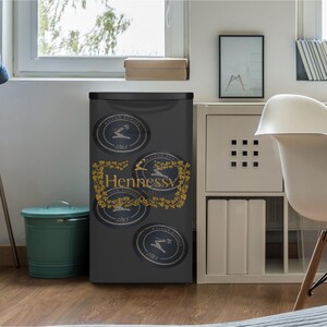 Puede incluir: Un minirefrigerador negro compacto con un patr&oacute;n repetitivo de logotipos circulares y el texto dorado Hennessy Maison Fond&eacute;e 1765. La puerta frontal est&aacute; decorada con un borde detallado de vides doradas que rodea la marca y emblemas.