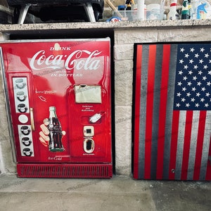 May include: Two red refrigerators side-by-side. The left refrigerator has a Coca-Cola logo and the text "Drink Coca-Cola in Bottles". The right refrigerator has an American flag design.