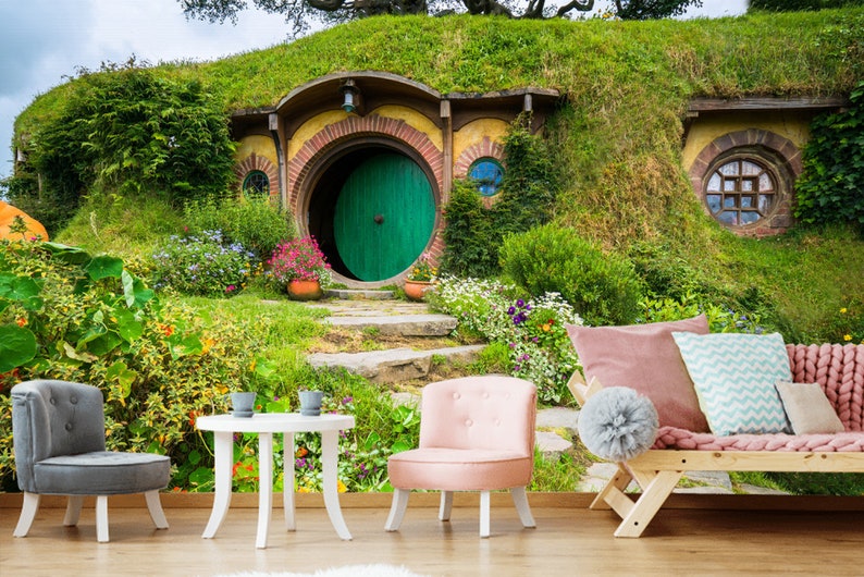 Pode incluir: Uma porta verde com um arco redondo leva a uma toca de hobbit, uma pequena casa redonda constru&iacute;da em uma colina gramada. A casa tem um exterior de tijolo e um telhado de palha. H&aacute; flores e plantas crescendo ao redor da casa.