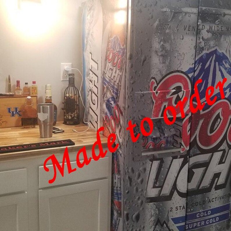 Puede incluir: Un refrigerador con un gr&aacute;fico de Coors Light que presenta un dise&ntilde;o de monta&ntilde;a y gotas de agua. Al lado hay una barra de madera con un vaso de acero inoxidable, varias botellas de licor de vidrio y una alfombrilla de bar negra sobre un mueble.