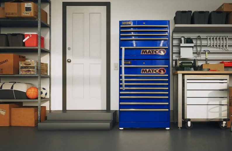 May include: A blue metal tool chest with multiple drawers. The chest has the word "MATCO" printed on it in white letters. The chest is sitting in a garage with a white door and a metal shelving unit.
