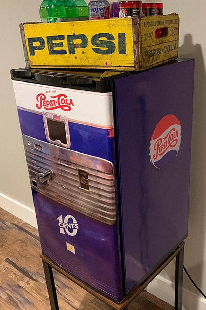 May include: A purple mini fridge with a vintage Pepsi-Cola design. The fridge has a metal coin slot and a sign that says "10 CENTS". The fridge is sitting on a wooden stand with black metal legs. A yellow wooden crate with the word "PEPSI" printed on it sits on top of the fridge.