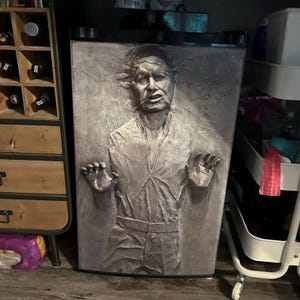 Può includere: Un mini frigorifero rettangolare con una scultura in rilievo tridimensionale e testurizzata sulla porta anteriore. La scultura raffigura una figura umana con le mani sollevate, caratterizzata da una finitura dettagliata, usurata e monocromatica.