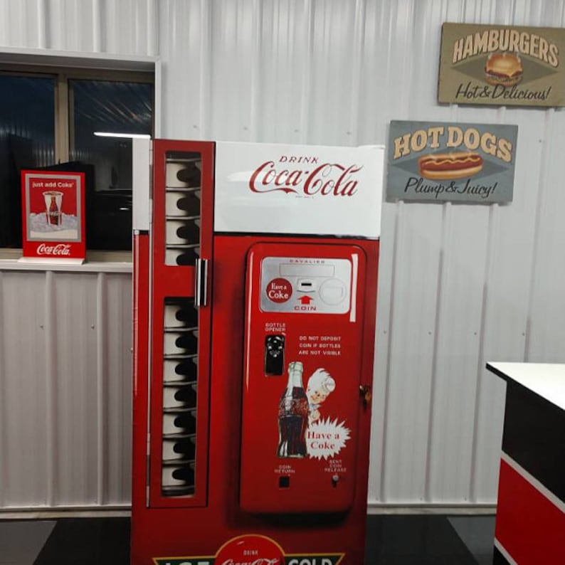May include: A vintage red Coca-Cola vending machine with a white top and a coin slot. The machine has a sign that says "Drink Coca-Cola" and a smaller sign that says "Have a Coke". The machine is in front of a white wall with signs for hamburgers, hot dogs, and Coca-Cola.
