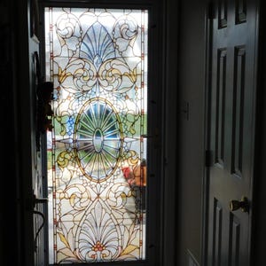 Op de afbeelding: Een gebrandschilderd glazen deurpaneel met een sierlijk ontwerp met bloemen en geometrische patronen in blauw, geel en rood. De deur is wit omlijst en staat gedeeltelijk open, waardoor een glimp van de buitenkant te zien is.