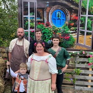 Pode incluir: Uma fam&iacute;lia de cinco pessoas vestidas com fantasias posam em frente a um cen&aacute;rio que retrata uma casa hobbit com uma porta redonda azul. O pai usa um avental marrom e segura um cajado. A m&atilde;e usa um vestido longo verde e marrom com padr&atilde;o floral. As tr&ecirc;s crian&ccedil;as usam v&aacute;rias fantasias.