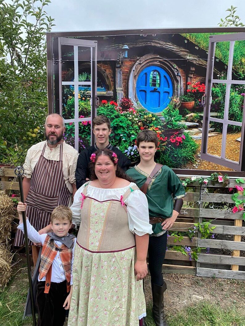 Pode incluir: Uma fam&iacute;lia de cinco pessoas vestidas com fantasias posam em frente a um cen&aacute;rio que retrata uma casa hobbit com uma porta redonda azul. O pai usa um avental marrom e segura um cajado. A m&atilde;e usa um vestido longo verde e marrom com padr&atilde;o floral. As tr&ecirc;s crian&ccedil;as usam v&aacute;rias fantasias.