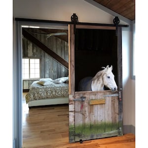 Può includere: Un cavallo bianco guarda fuori da una porta di stalla in legno con una porta di fienile rustica che scorre per rivelare una camera da letto con un letto e una finestra.