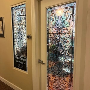May include: A white door with a stained glass window featuring a colorful floral design. The door has a silver door handle. A smaller window to the left has a sign that reads "Hair Design by Nancy".