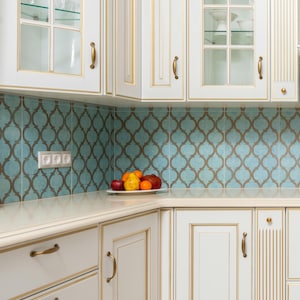 May include: A corner kitchen cabinet set with white doors, gold trim, and brass handles. The backsplash features a teal and brown patterned tile. A plate of fruit sits on the countertop. The cabinets have glass-fronted upper sections.