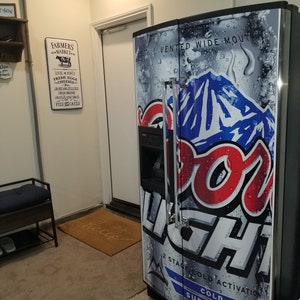 May include: A stainless steel refrigerator with a Coors Light can design on the door. The refrigerator has a water and ice dispenser and a vented wide mouth.