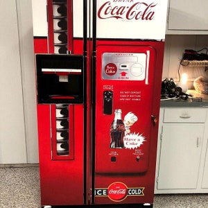 May include: A red Coca-Cola refrigerator with a vintage vending machine design. The refrigerator has a bottle opener, coin return, and a bent coin release. The refrigerator is labeled "Have a Coke" and "ICE COLD".