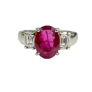 以下が含まれることがあります： 大きな楕円形のルビーと、両側に2つの小さなバゲットカットダイヤモンドがセットされたプラチナリングです。