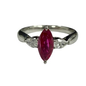 May include: A silver-toned ring featuring a central marquise-cut vibrant red gemstone flanked by two smaller pear-cut clear white gemstones. The band is smooth and polished, providing a clean setting for the three-stone arrangement.