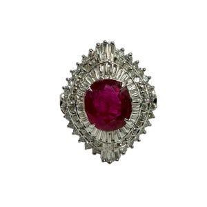 Może przedstawiać: Srebrny pierścionek z dużym, owalnym rubinem w centrum, otoczonym mniejszymi diamentami o szlifie bagietowym. Projekt pierścionka jest symetryczny, a głęboki czerwony kolor rubinu kontrastuje z jasną bielą diamentów i srebrnej oprawy.