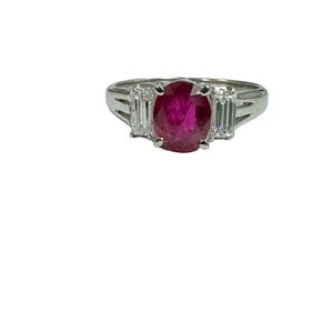 May include: A platinum ring with a large oval ruby gemstone and two smaller baguette diamonds on either side.