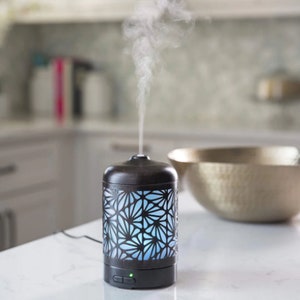 May include: A black essential oil diffuser with a floral pattern and a blue light. The diffuser is on a white countertop and is emitting steam.